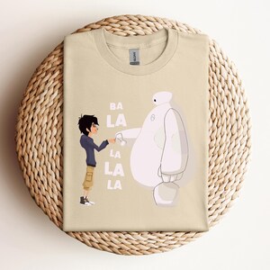 K&ouml;nnte beinhalten: Ein beiges T-Shirt mit einem Grafikdruck von zwei Figuren aus dem Animationsfilm Big Hero 6. Die Figuren sind Hiro Hamada und Baymax. Der Text "BA LA LA LA LA" ist auf dem T-Shirt gedruckt.