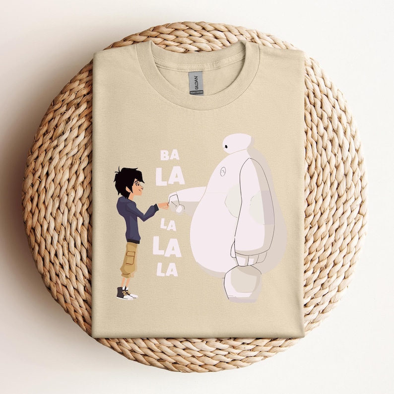 K&ouml;nnte beinhalten: Ein beiges T-Shirt mit einem Grafikdruck von zwei Figuren aus dem Animationsfilm Big Hero 6. Die Figuren sind Hiro Hamada und Baymax. Der Text "BA LA LA LA LA" ist auf dem T-Shirt gedruckt.
