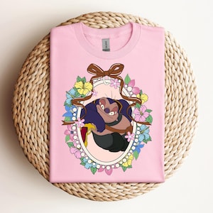 Puede incluir: Una camiseta rosa claro con un personaje de dibujos animados dentro de un marco ovalado. El marco est&aacute; decorado con flores y una cinta marr&oacute;n. El personaje es una criatura de color p&uacute;rpura oscuro y marr&oacute;n con una expresi&oacute;n astuta.