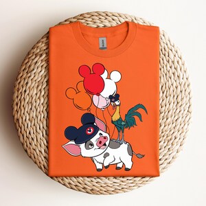 Op de afbeelding: Oranje t-shirt met een cartoon illustratie van een varken met een hoed, een haan en Mickey Mouse-vormige ballonnen in rood, wit en roze. Het shirt is op een geweven oppervlak gevouwen.