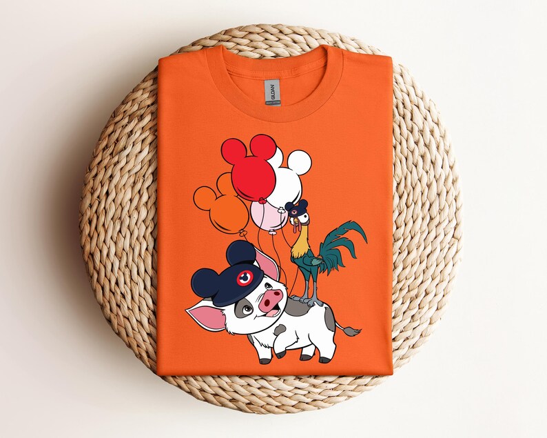 Op de afbeelding: Oranje t-shirt met een cartoon illustratie van een varken met een hoed, een haan en Mickey Mouse-vormige ballonnen in rood, wit en roze. Het shirt is op een geweven oppervlak gevouwen.