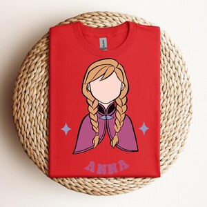 May include: A bright red t-shirt featuring a stylised graphic of a character with blonde hair in two braids, wearing a purple cape. The character has no facial features. Below the graphic, the name ANNA is printed in a purple, wavy font.
