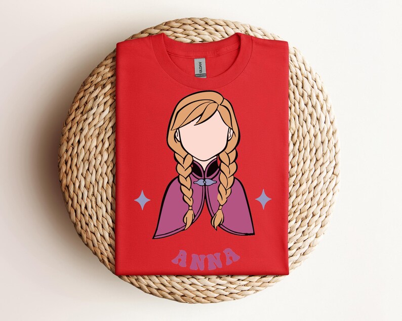May include: A bright red t-shirt featuring a stylised graphic of a character with blonde hair in two braids, wearing a purple cape. The character has no facial features. Below the graphic, the name ANNA is printed in a purple, wavy font.