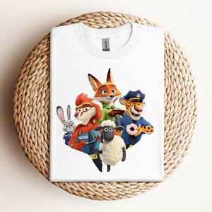 May include: White t-shirt featuring a colorful graphic of characters from the animated movie Zootopia. The design includes a fox, a bunny, a sheep, and a cheetah, with a donut. The shirt is displayed on a woven surface.