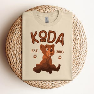 May include: A beige t-shirt with a cartoon brown bear and the text "KODA EST. 2003".