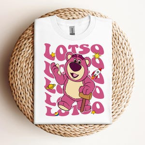 Puede incluir: Camiseta blanca con un oso de dibujos animados rosa sosteniendo un martillo. El oso tiene la nariz morada y los ojos marrones. El texto "LOTSO" se repite en un patr&oacute;n de espiral rosa y blanco alrededor del oso.