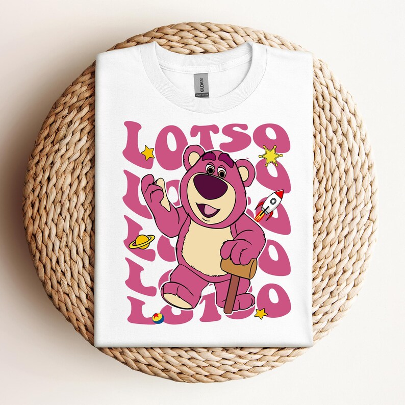 Puede incluir: Camiseta blanca con un oso de dibujos animados rosa sosteniendo un martillo. El oso tiene la nariz morada y los ojos marrones. El texto "LOTSO" se repite en un patr&oacute;n de espiral rosa y blanco alrededor del oso.