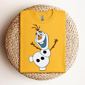 Pode incluir: Uma camiseta amarela com uma imagem de desenho animado de Olaf, o boneco de neve do filme Frozen, com um grande sorriso e os bra&ccedil;os estendidos.