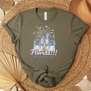 Puede incluir: Camiseta verde oliva con una ilustraci&oacute;n de un castillo, fuegos artificiales y el texto "Florida!!! We're hell of a drug."