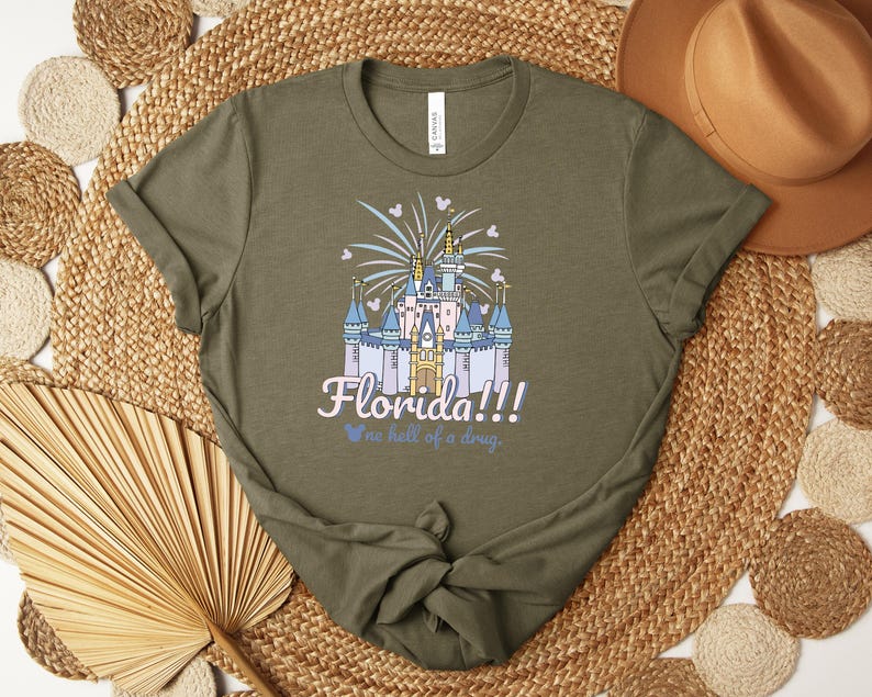 Puede incluir: Camiseta verde oliva con una ilustraci&oacute;n de un castillo, fuegos artificiales y el texto "Florida!!! We're hell of a drug."