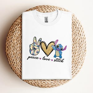 以下が含まれることがあります： リロ＆スティッチのスティッチが描かれた白いTシャツ。ピースサイン、ハート、そして「peace love stitch」の文字が書かれています。