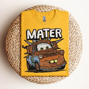 May include: Mustard yellow t-shirt featuring a graphic of Mater from the movie Cars. The word "MATER" is printed in large, white, block letters above the cartoon image of the character. The shirt is folded on a woven, circular surface.
