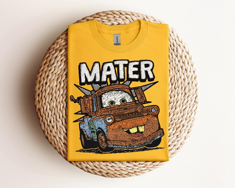 May include: Mustard yellow t-shirt featuring a graphic of Mater from the movie Cars. The word "MATER" is printed in large, white, block letters above the cartoon image of the character. The shirt is folded on a woven, circular surface.