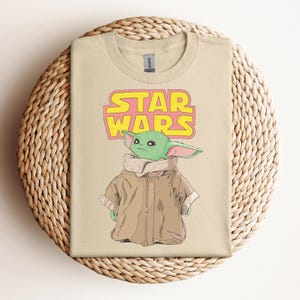 Peut inclure: T-shirt beige avec un dessin d'un personnage extraterrestre vert de Star Wars, avec les mots "STAR WARS" en jaune et rose. L'extraterrestre porte un manteau marron. Le t-shirt est sur un panier tress&eacute;.