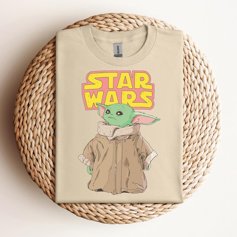 Peut inclure: T-shirt beige avec un dessin d'un personnage extraterrestre vert de Star Wars, avec les mots "STAR WARS" en jaune et rose. L'extraterrestre porte un manteau marron. Le t-shirt est sur un panier tress&eacute;.