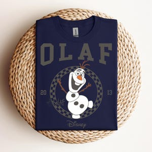 Może przedstawiać: Koszulka w kolorze granatowym z grafiką Olafa z Krainy Lodu. Grafika przedstawia Olafa w kwadratowym k&oacute;łku z napisem "Olaf" nad nim i "Disney" pod nim.