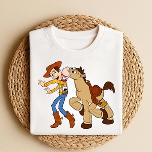 Peut inclure: T-shirt blanc avec une illustration de Woody de Toy Story et un cheval. Woody porte un chapeau de cowboy, une chemise jaune, un jean bleu et des bottes marron. Le cheval est marron avec une selle.