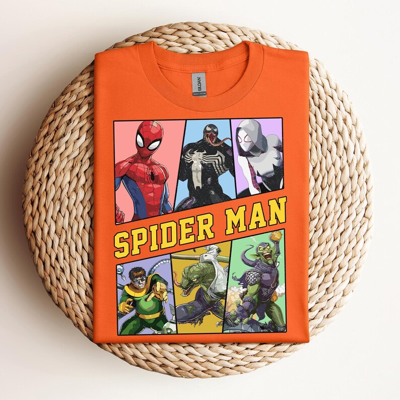 Pode incluir: Camiseta laranja com um gr&aacute;fico de banda desenhada do Homem-Aranha e outros personagens. As palavras "SPIDER MAN" est&atilde;o em uma fonte amarela e preta. A camiseta est&aacute; dobrada em uma superf&iacute;cie circular tecida.