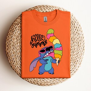 May include: Orange t-shirt featuring a cartoon character wearing sunglasses and holding a large ice cream cone. The text "hello summer" is written in black script. The shirt is folded on a woven, circular surface.