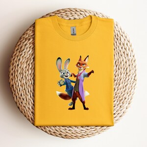 May include: Mustard yellow t-shirt featuring a cartoon image of a fox and a rabbit. The fox is wearing a purple coat and the rabbit is wearing a blue jacket. The t-shirt is folded on a woven, circular surface.