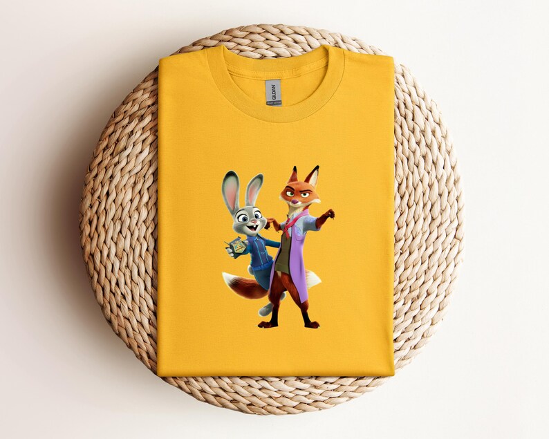 May include: Mustard yellow t-shirt featuring a cartoon image of a fox and a rabbit. The fox is wearing a purple coat and the rabbit is wearing a blue jacket. The t-shirt is folded on a woven, circular surface.