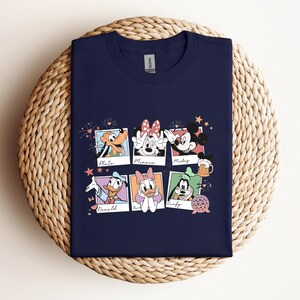May include: A navy blue t-shirt featuring a graphic print of six polaroid-style frames. Each frame contains a character illustration with their name written below: Pluto, Minnie, Mickey, Donald, Daisy, and Goofy. Small icons like fireworks, a beer mug, and a geometric sphere are scattered around the frames.