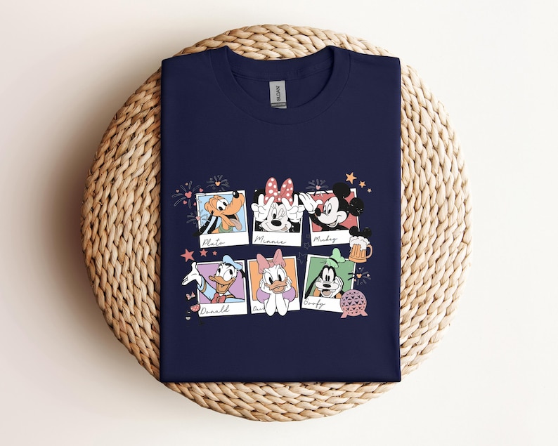 May include: A navy blue t-shirt featuring a graphic print of six polaroid-style frames. Each frame contains a character illustration with their name written below: Pluto, Minnie, Mickey, Donald, Daisy, and Goofy. Small icons like fireworks, a beer mug, and a geometric sphere are scattered around the frames.