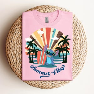 May include: Pale pink t-shirt with a cartoon character wearing sunglasses and holding a surfboard, set against a retro sunset design with palm trees. The text "Summer Vibes" is written in a script font below the graphic.