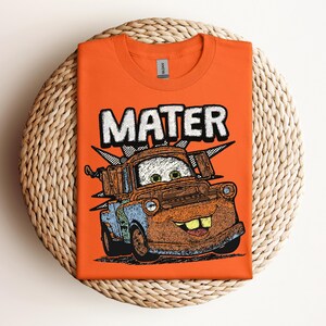 May include: Orange t-shirt featuring a graphic of Mater from the movie Cars. The word "MATER" is printed above the character in white block letters. The cartoon image of Mater is in shades of brown, blue, and green.