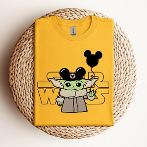 Puede incluir: Una camiseta amarilla con un personaje verde que lleva un sombrero de Mickey Mouse y sostiene un globo negro de Mickey Mouse. El texto "STAR WARS" est&aacute; impreso en la camiseta.