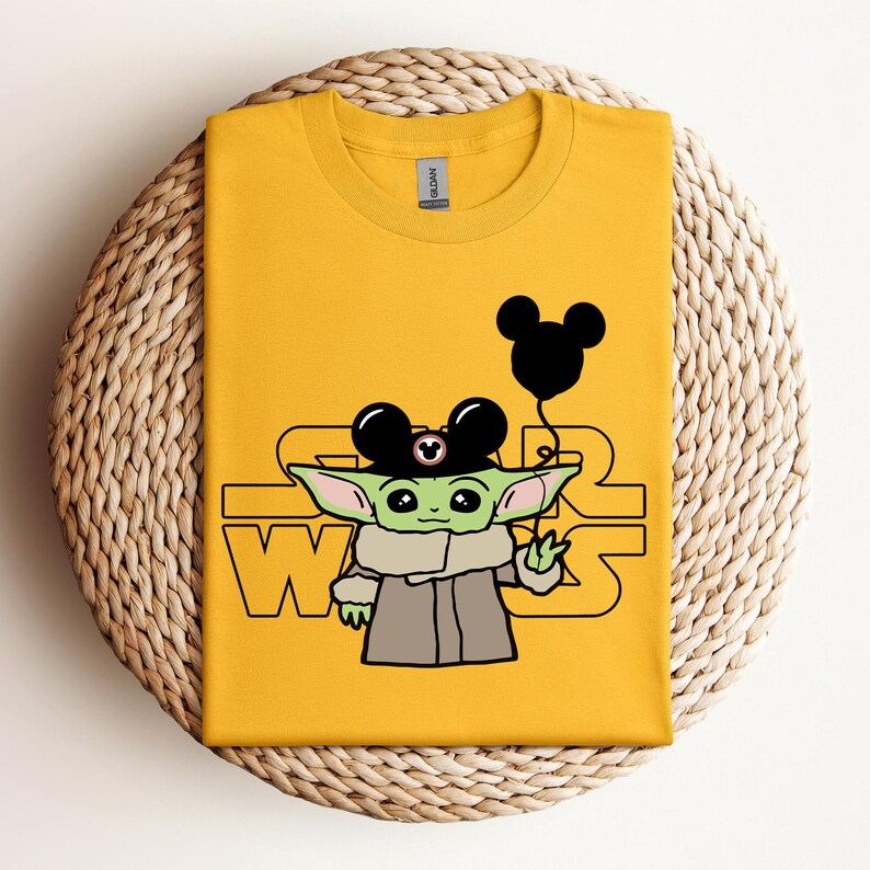 Puede incluir: Una camiseta amarilla con un personaje verde que lleva un sombrero de Mickey Mouse y sostiene un globo negro de Mickey Mouse. El texto "STAR WARS" est&aacute; impreso en la camiseta.
