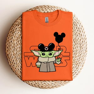Puede incluir: Camiseta naranja con un personaje alien&iacute;gena verde que lleva un sombrero de Mickey Mouse y sostiene un globo de Mickey Mouse. El texto "Star Wars" est&aacute; impreso en la camiseta.
