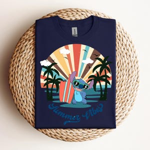 May include: Navy blue t-shirt with a cartoon character wearing sunglasses and holding a surfboard, set against a sunset scene with palm trees. The text "Summer Vibes" is written in a script font. The shirt is on a woven surface.