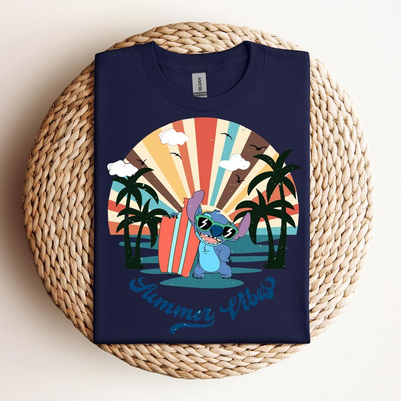May include: Navy blue t-shirt with a cartoon character wearing sunglasses and holding a surfboard, set against a sunset scene with palm trees. The text "Summer Vibes" is written in a script font. The shirt is on a woven surface.