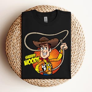 May include: Black t-shirt featuring a cartoon illustration of Sheriff Woody from Toy Story. The character is depicted in a cowboy hat, yellow shirt, red neckerchief, and holding a lasso. The text "SHERIFF WOODY" is displayed below the character.
