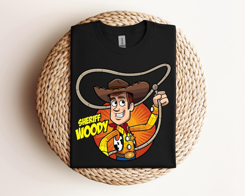 May include: Black t-shirt featuring a cartoon illustration of Sheriff Woody from Toy Story. The character is depicted in a cowboy hat, yellow shirt, red neckerchief, and holding a lasso. The text "SHERIFF WOODY" is displayed below the character.