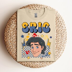 May include: A tan t-shirt featuring the name "ERIC" in large, retro-style lettering with blue and yellow accents. Below the name is a cartoon illustration of a man with blue eyes and black hair. The design includes stars, lightning bolts, and paint splatters.
