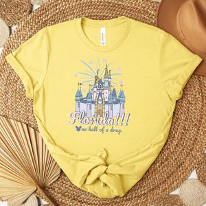 Puede incluir: Una camiseta amarilla con un gr&aacute;fico de un castillo de cuento de hadas, el texto "Florida!!!" y "one hell of a drug." El castillo es azul, morado y dorado, con fuegos artificiales. La camiseta est&aacute; hecha de un material suave. Es una prenda casual.