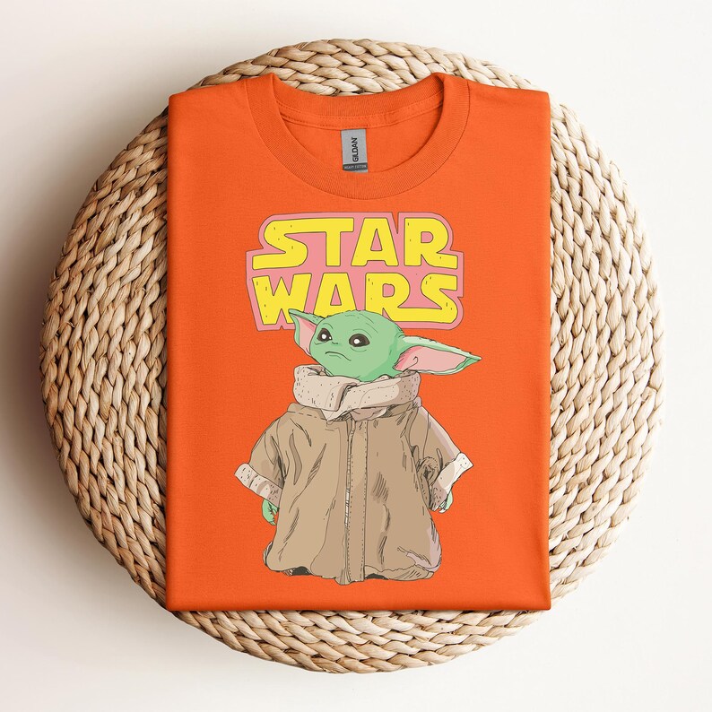 Peut inclure: T-shirt orange avec un motif de Grogu de Star Wars, et les mots "STAR WARS" en jaune et rose. Grogu porte son manteau marron. Le t-shirt est pr&eacute;sent&eacute; sur une surface circulaire tiss&eacute;e.