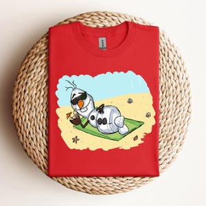 Op de afbeelding: Rood T-shirt met een cartoon afbeelding van Olaf uit Frozen, die een zonnebril draagt en ontspant op een strandhanddoek met een tropisch drankje.