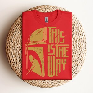 May include: Red t-shirt with a gold graphic of a helmet and the text "THIS IS THE WAY." The shirt is folded and placed on a woven, circular surface. The design has a distressed, vintage look.