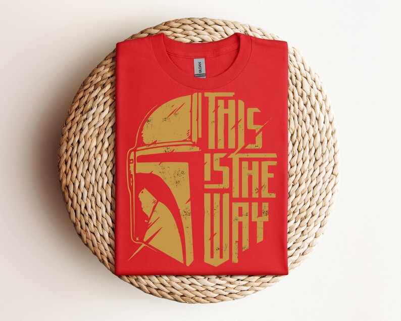 May include: Red t-shirt with a gold graphic of a helmet and the text "THIS IS THE WAY." The shirt is folded and placed on a woven, circular surface. The design has a distressed, vintage look.