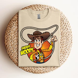May include: A tan t-shirt featuring a cartoon illustration of Sheriff Woody from Toy Story. The character is depicted with a brown cowboy hat, yellow shirt, red neckerchief, and a lasso. The words "SHERIFF WOODY" are printed below the character.