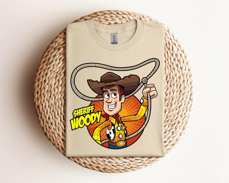 May include: A tan t-shirt featuring a cartoon illustration of Sheriff Woody from Toy Story. The character is depicted with a brown cowboy hat, yellow shirt, red neckerchief, and a lasso. The words "SHERIFF WOODY" are printed below the character.