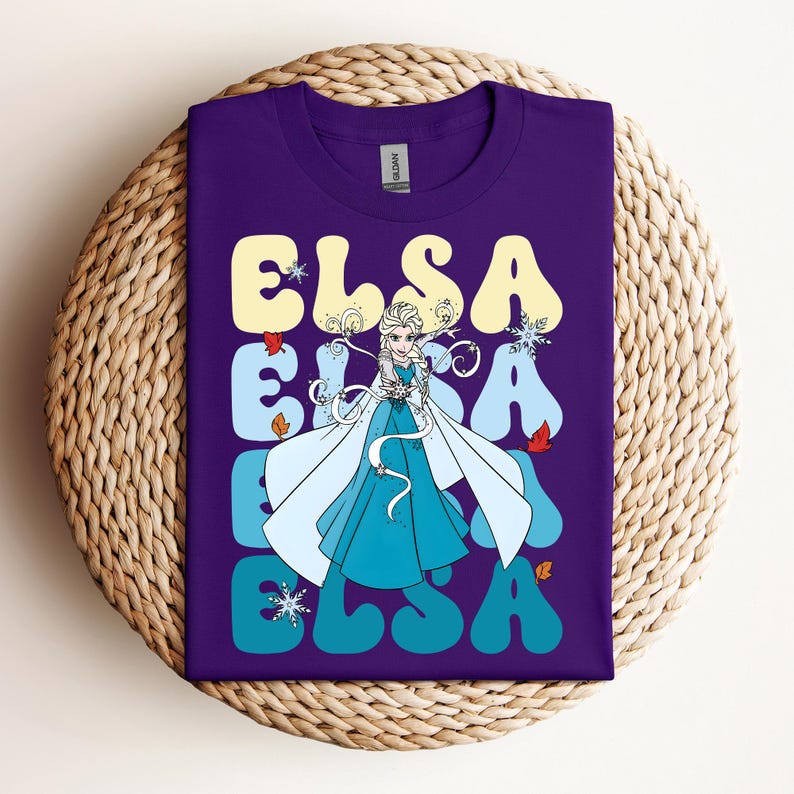 Op de afbeelding: Een paars t-shirt met het woord "ELSA" in grote, gele letters in retrostijl. Het ontwerp bevat een illustratie van Elsa uit Frozen in een blauwe jurk en cape, met sneeuwvlokken en herfstbladeren.