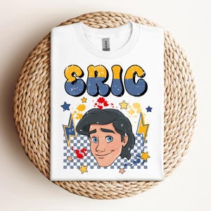May include: White t-shirt featuring the name "ERIC" in yellow and blue retro font. Below the name is a cartoon illustration of a man's face with blue eyes and black hair. The design includes stars, lightning bolts, and a checkered pattern.