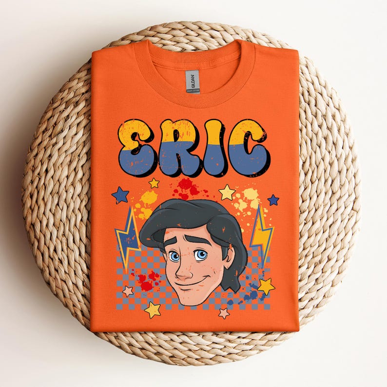 May include: Orange t-shirt with the name "ERIC" in large, retro-style lettering. The shirt features a cartoon illustration of a character with black hair and blue eyes, surrounded by stars, lightning bolts, and paint splatters.