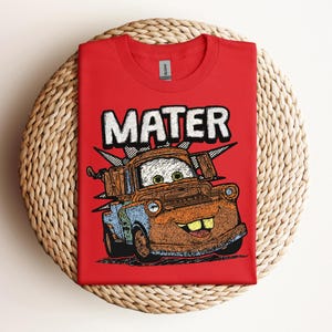 May include: A red t-shirt featuring a cartoon image of Mater from the movie Cars. The word "MATER" is printed above the character in white block letters. The shirt is displayed on a woven, circular surface.