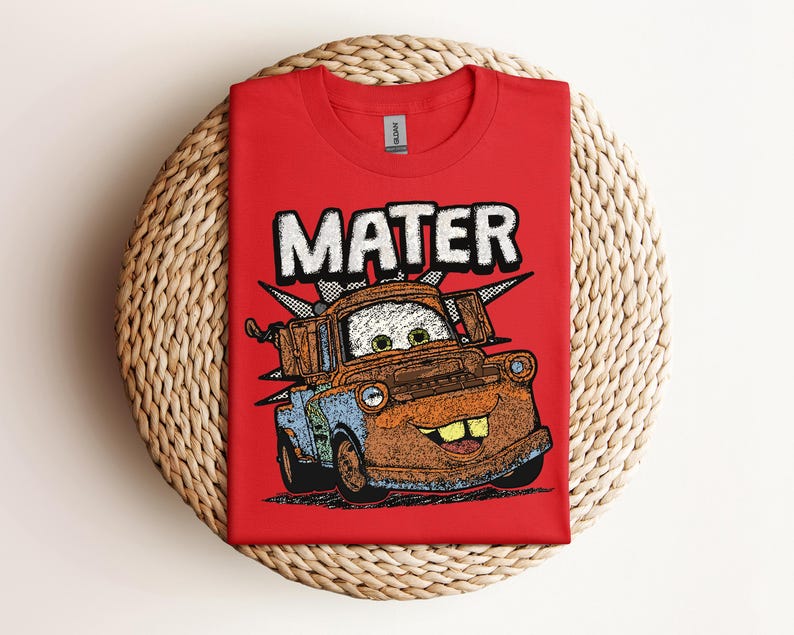 May include: A red t-shirt featuring a cartoon image of Mater from the movie Cars. The word "MATER" is printed above the character in white block letters. The shirt is displayed on a woven, circular surface.