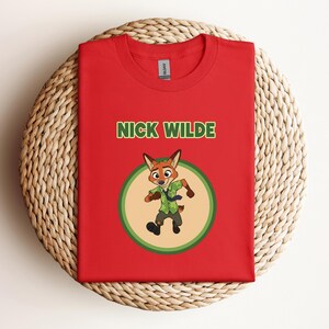 May include: Red t-shirt featuring the text "NICK WILDE" in green above a cartoon fox character in a green and tan circle. The fox is wearing a green shirt and tie. The shirt is displayed on a woven, circular surface.